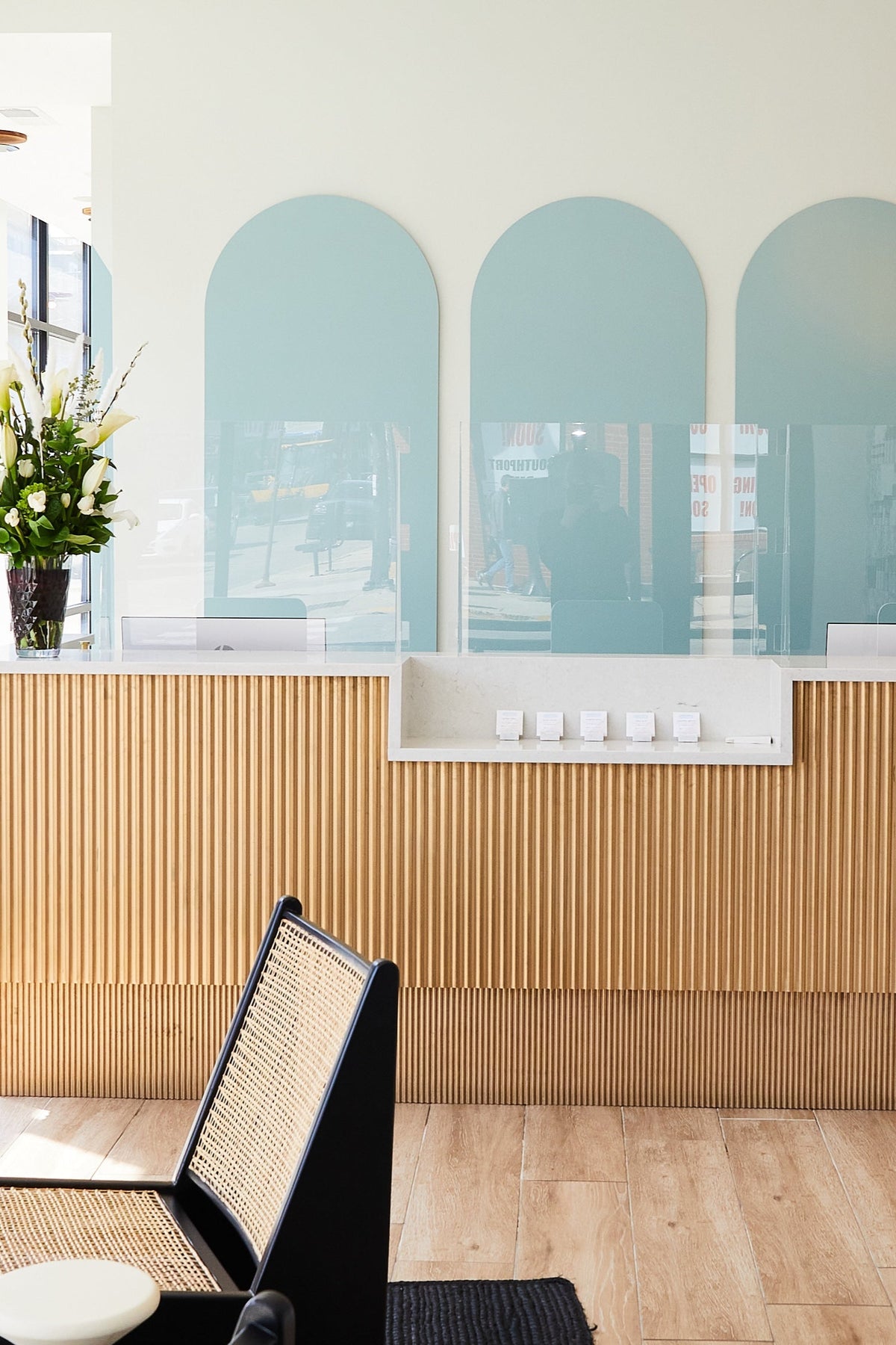 Reception area with a wooden front desk and decorative panels. A vase with flowers sits on the desk. Modern hanging lights and a patterned wall in the background. Large windows on the left.
