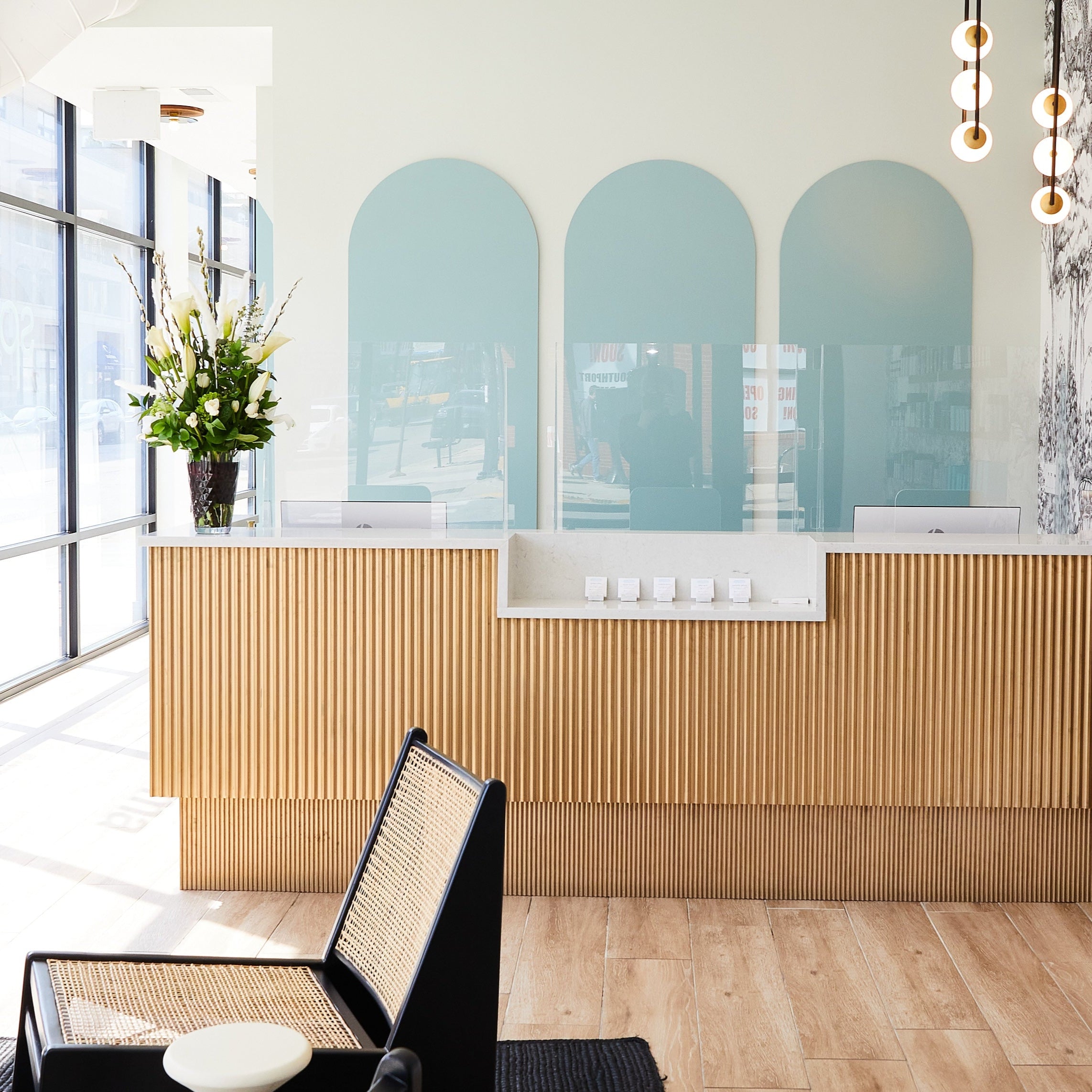 Reception area with a wooden front desk and decorative panels. A vase with flowers sits on the desk. Modern hanging lights and a patterned wall in the background. Large windows on the left.
