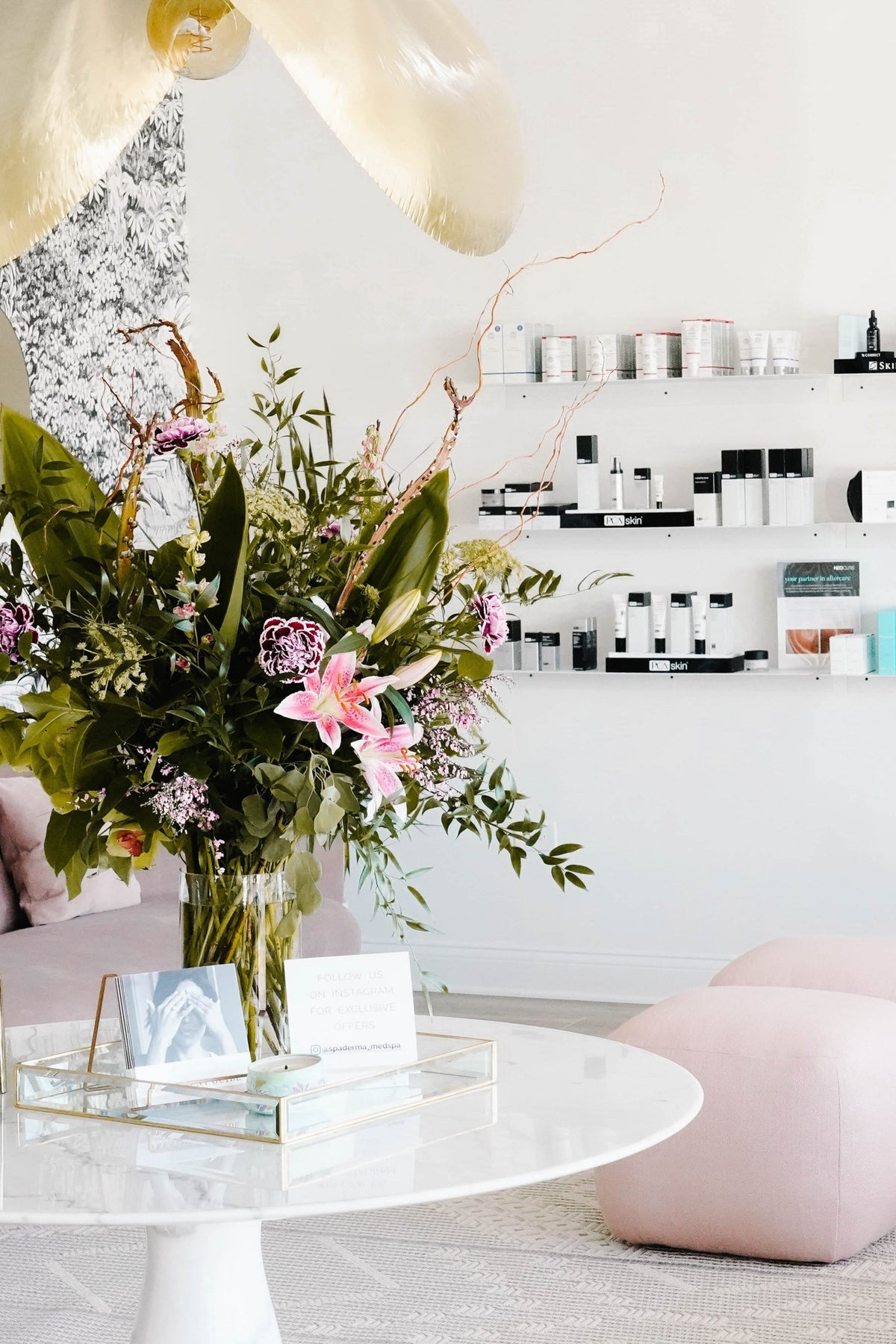 A chic, modern interior with a round table and floral arrangement. Shelves with products line the walls, and pink seating is arranged on the floor.