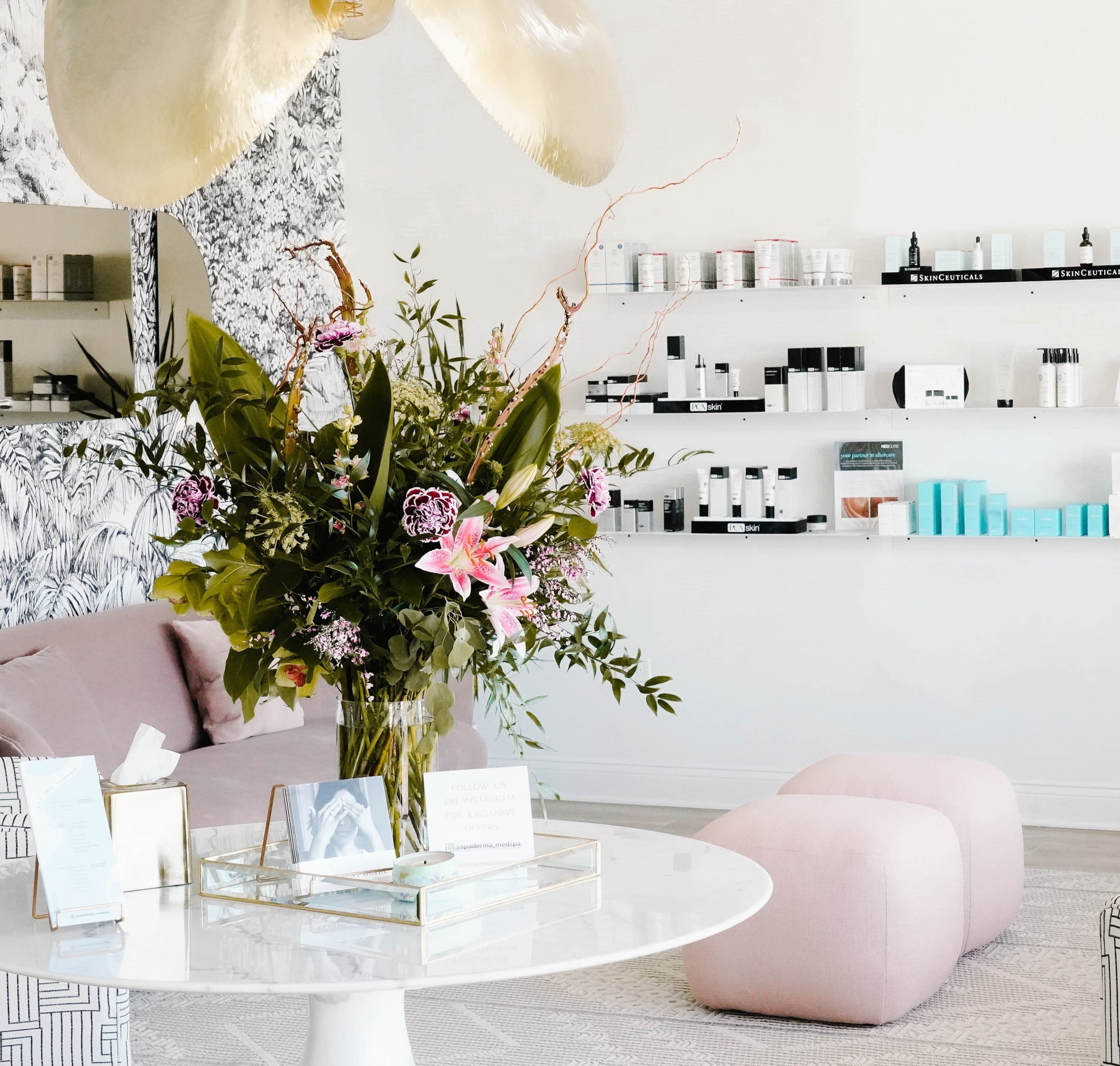 A chic, modern interior with a round table and floral arrangement. Shelves with products line the walls, and pink seating is arranged on the floor.
