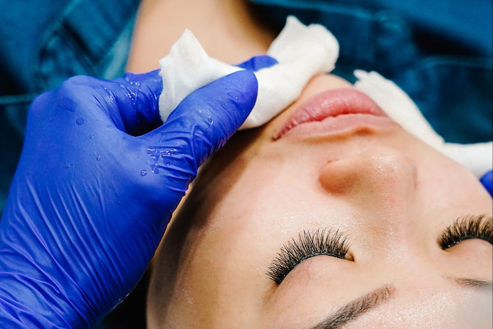 Person receiving a facial treatment, eyes closed, with blue-gloved hands holding a cloth on their face.