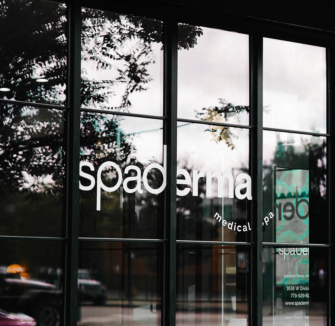 Reflective glass storefront with the word "spademia" and a partial view of a street and trees outside.