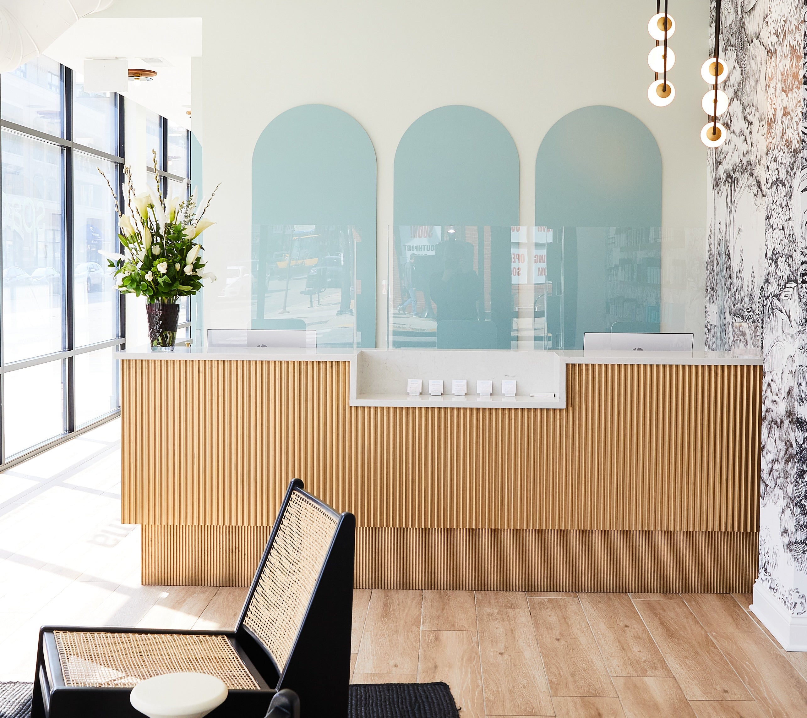 Reception area with a wooden front desk and decorative panels. A vase with flowers sits on the desk. Modern hanging lights and a patterned wall in the background. Large windows on the left.
