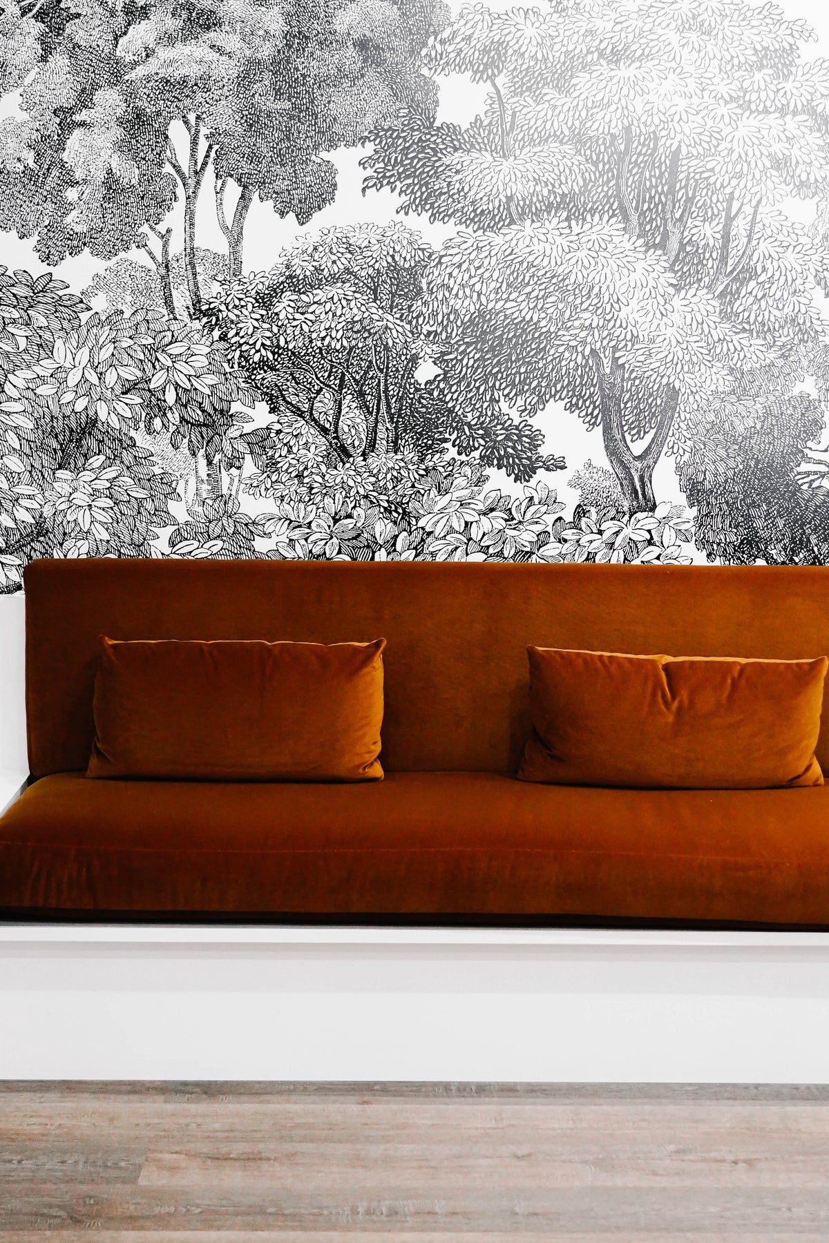 A modern seating area with rust-colored sofas and cushioned seats against a detailed black-and-white tree mural on the wall, set on a wooden floor.