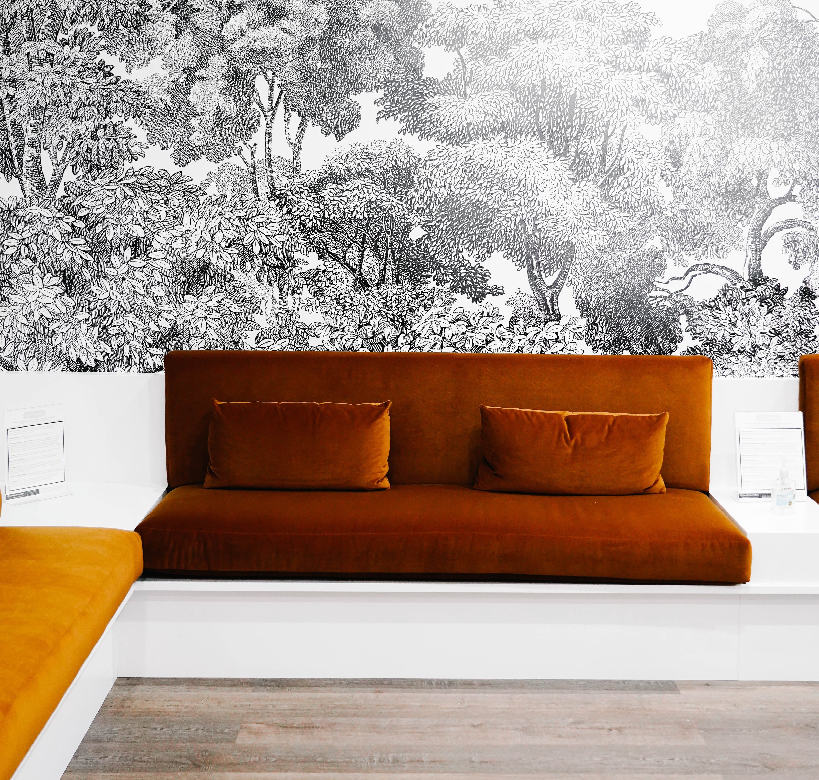 A modern seating area with rust-colored sofas and cushioned seats against a detailed black-and-white tree mural on the wall, set on a wooden floor.