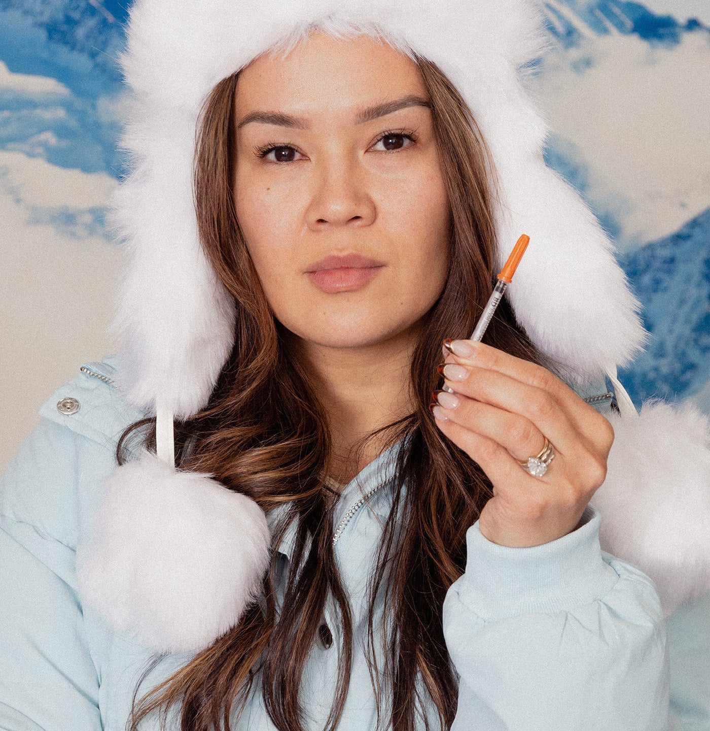 Woman in light blue coat with white fur trim holding a syringe against a colorful abstract background