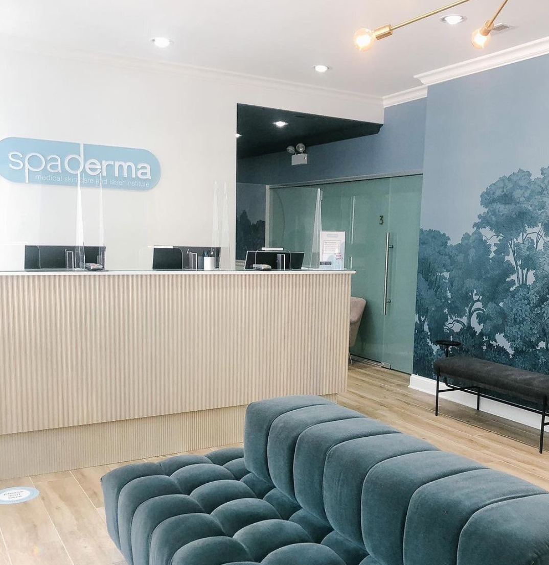 Modern reception area with a blue tufted sofa, light wood flooring, a reception desk with a "Spaderma" sign, and a wall mural of trees.