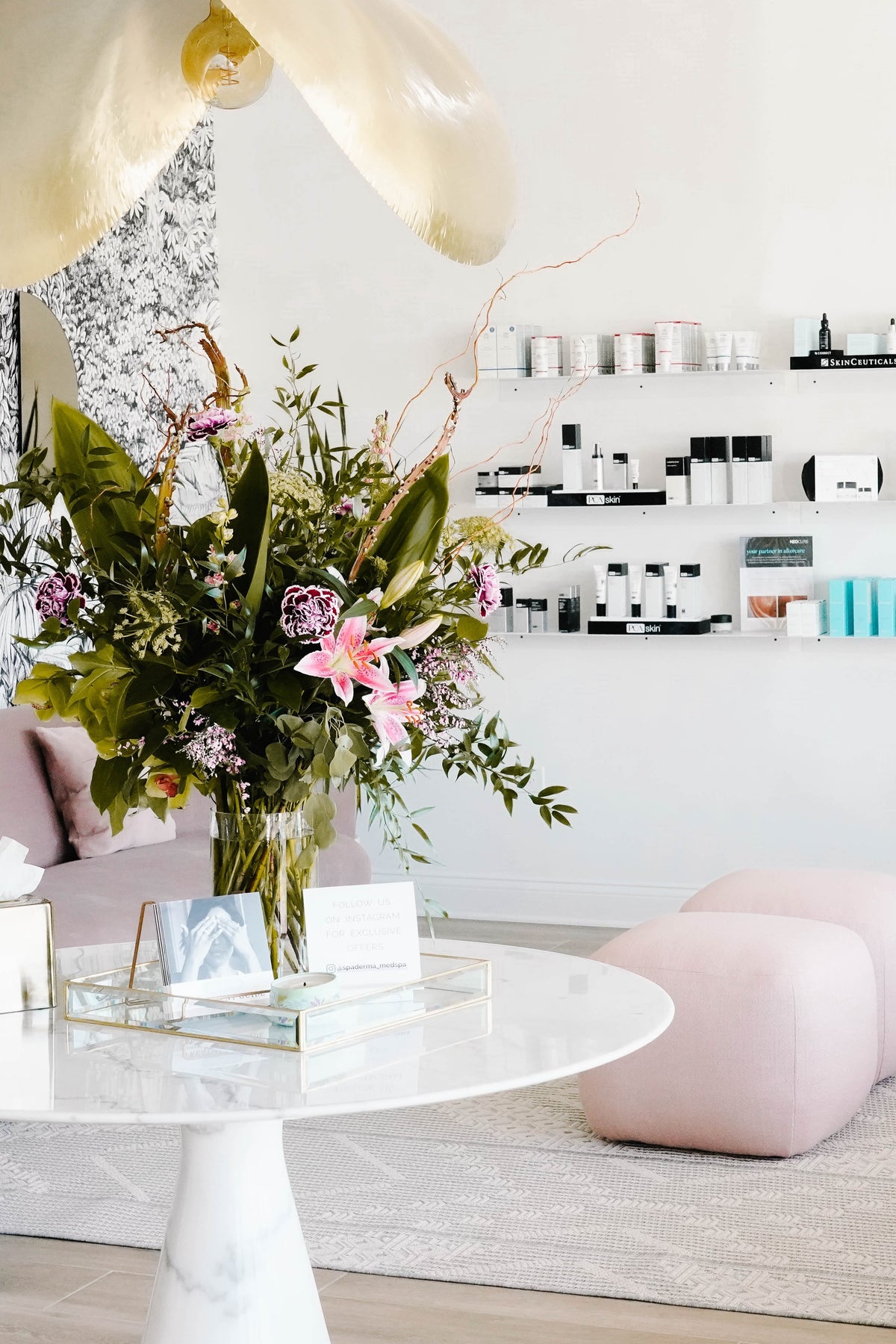 A chic, modern interior with a round table and floral arrangement. Shelves with products line the walls, and pink seating is arranged on the floor.