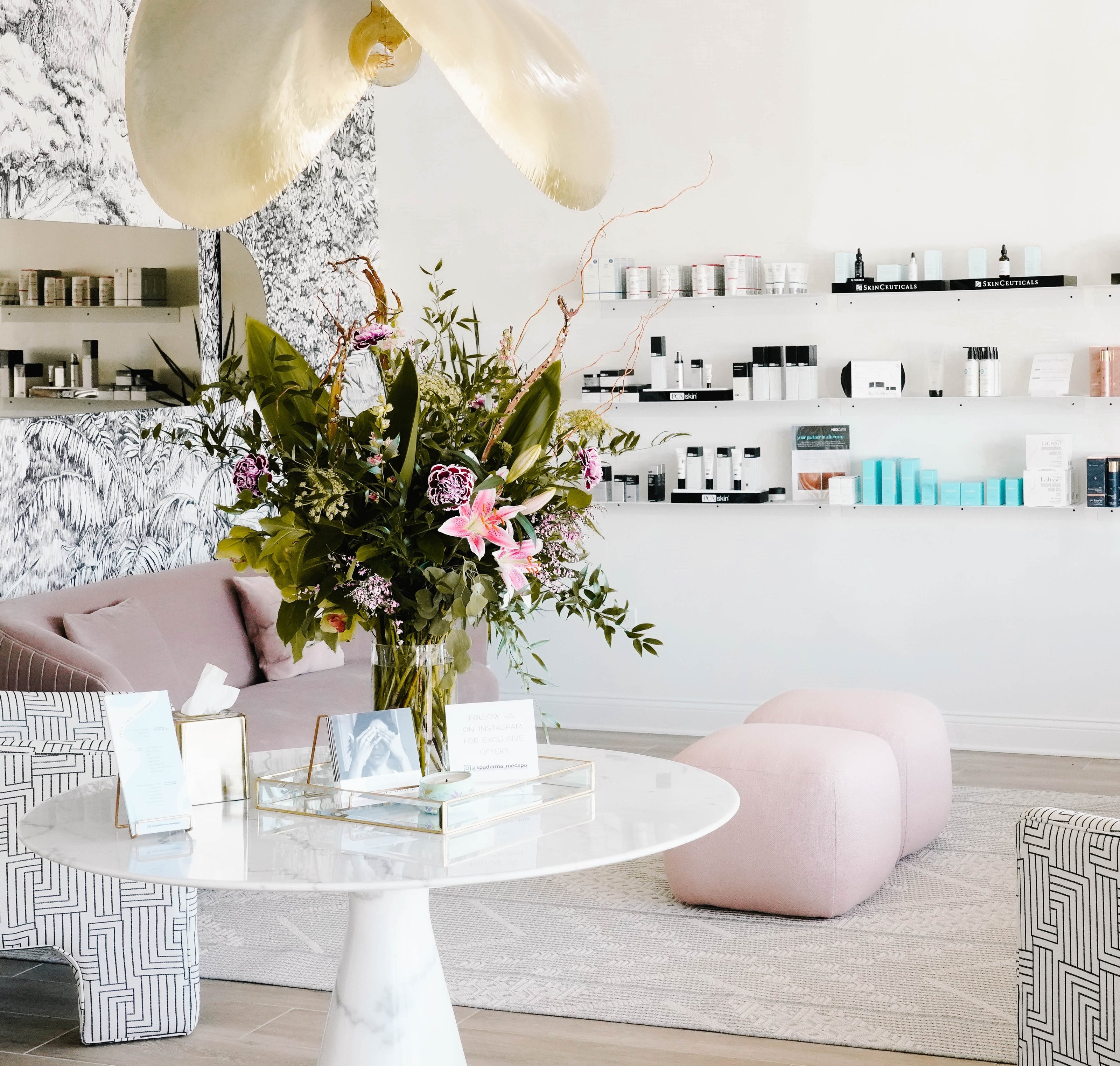 A chic, modern interior with a round table and floral arrangement. Shelves with products line the walls, and pink seating is arranged on the floor.